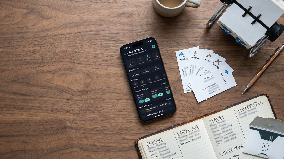 iPhone showing FlipPro AI's Black Book with 38 contacts categorised by type, on a walnut workbench beside a vintage cracked-leather notebook with handwritten PAINTERS, ELECTRICIANS, TRADIES sections, five tradie business cards fanned out and a Rolodex card holder